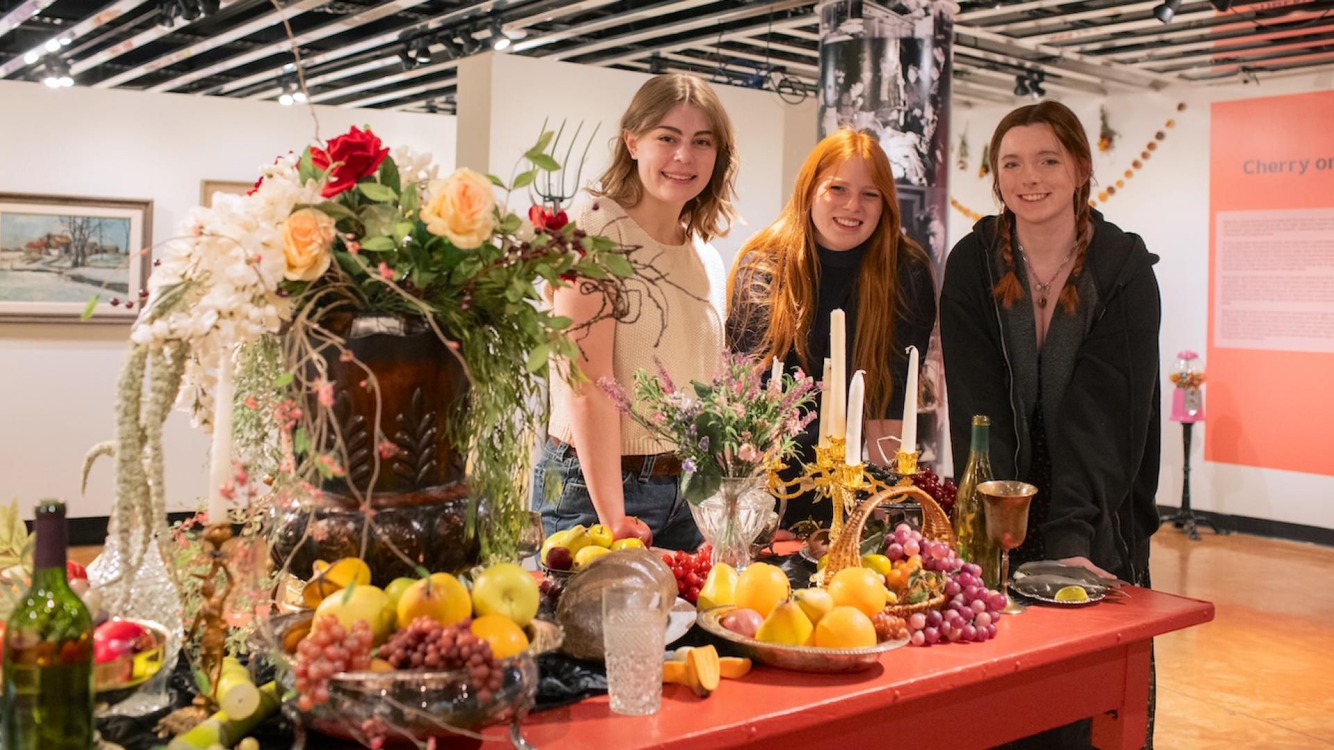 Three college-aged women stand around a staged opulent feast in an art gallery. 