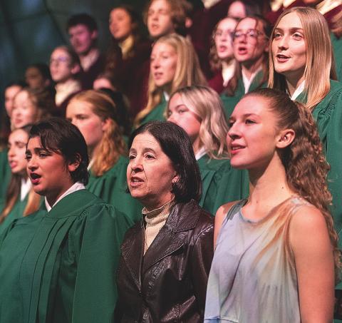 A mature woman stands with young people singing. 