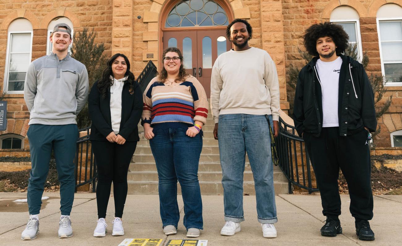 Five cool college students stand in front of a turn-of-the-century college building on the Gustavus Adolphus College campus