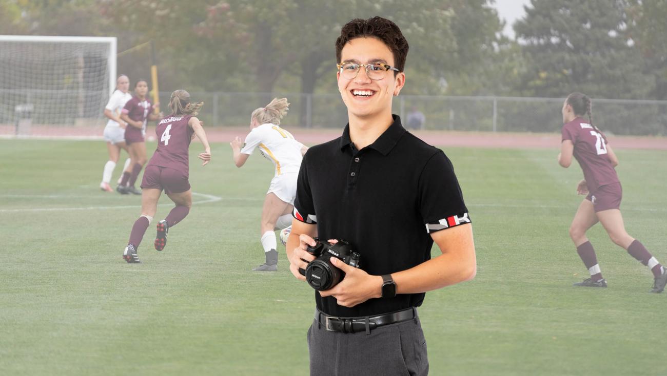 A young man smiling with a camera in his hands with a soccer game going on in the background. 