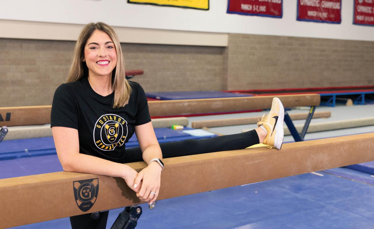 Aryn Bell DeGrood standing with one leg on the beam, in the Gustavus Sponberg Gymnastics studio 