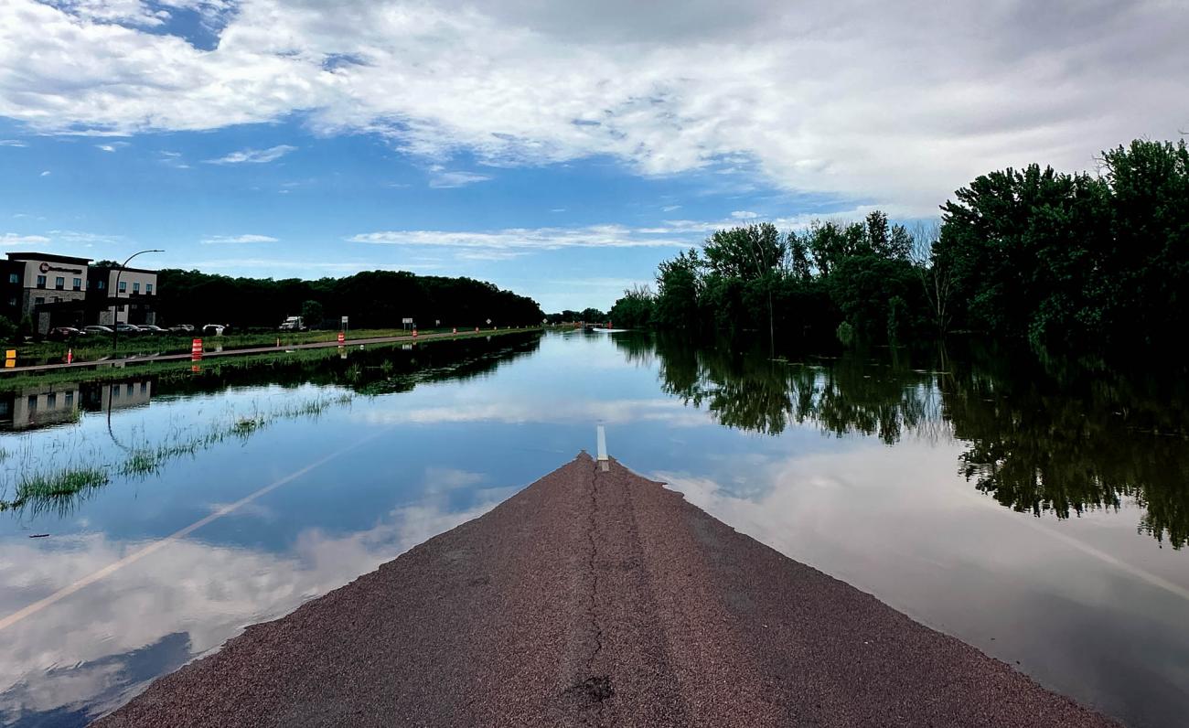 Water submerging a flooded highway reflects the sky and the trees around it.