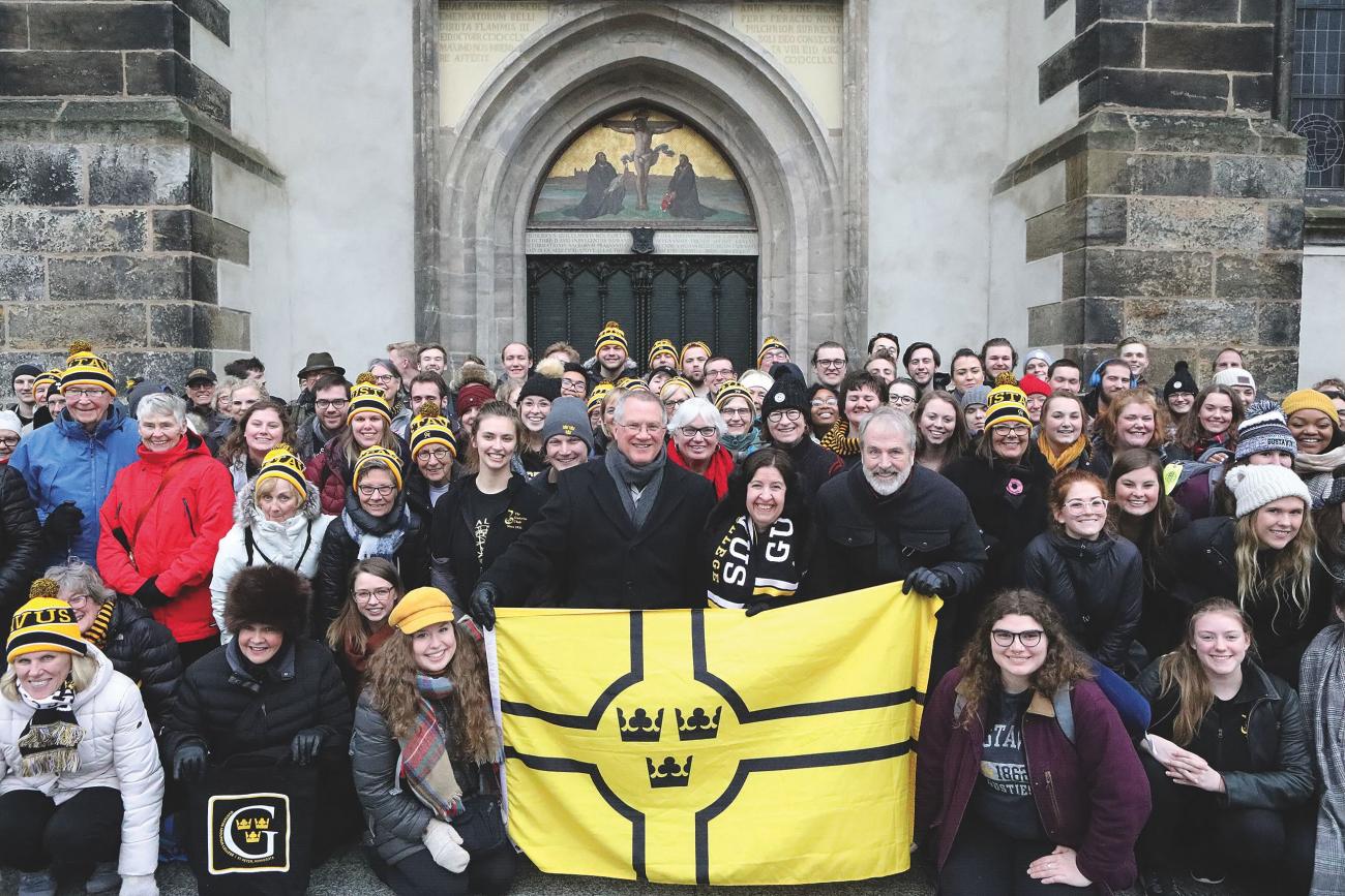  A large multigenerational collection of people with a Gustavus Adolphus flag stand outside church doors. 