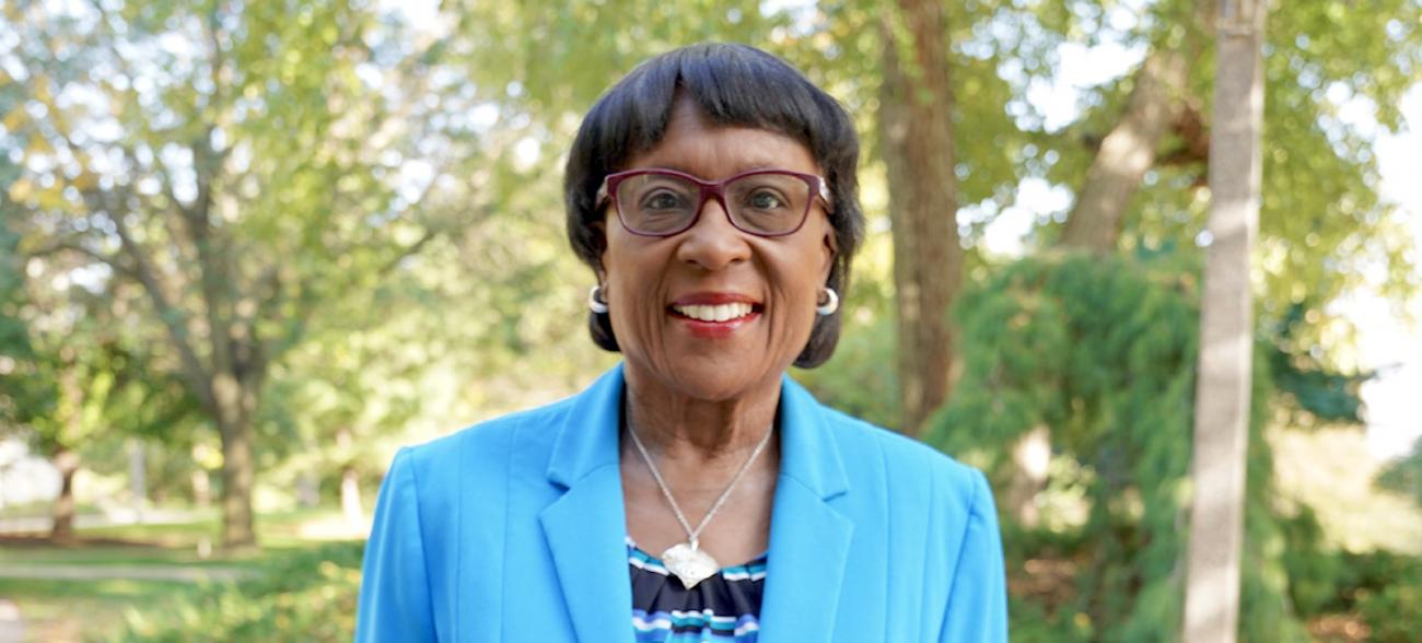 A distinguished woman stands in front of a park-like setting.