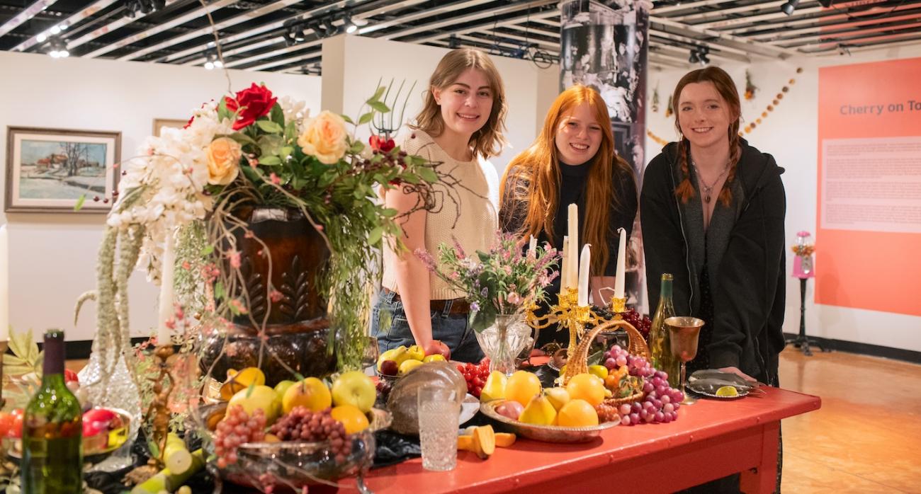 Three college-aged women stand around a staged opulent feast in an art gallery. 