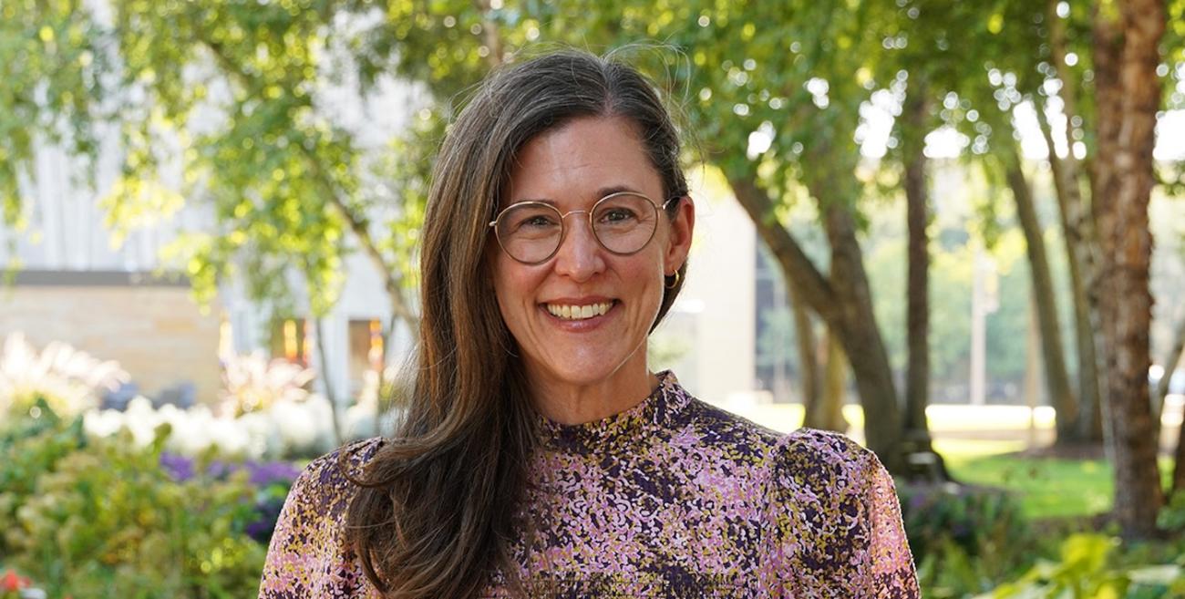 A woman with glasses stands and smiles in a park-like setting. 