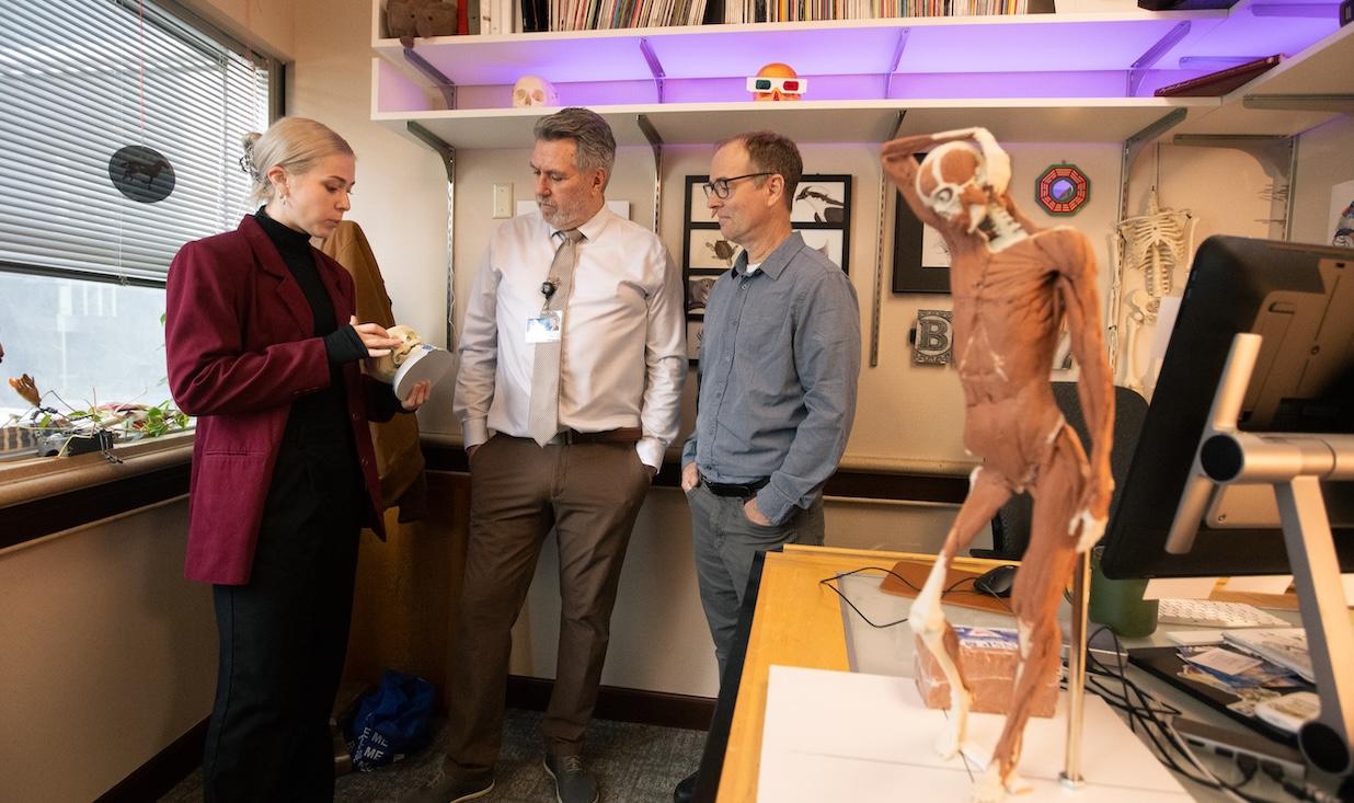 A woman shows two men a model of a skull in an office that also includes a model of the human body, complete with musculature.