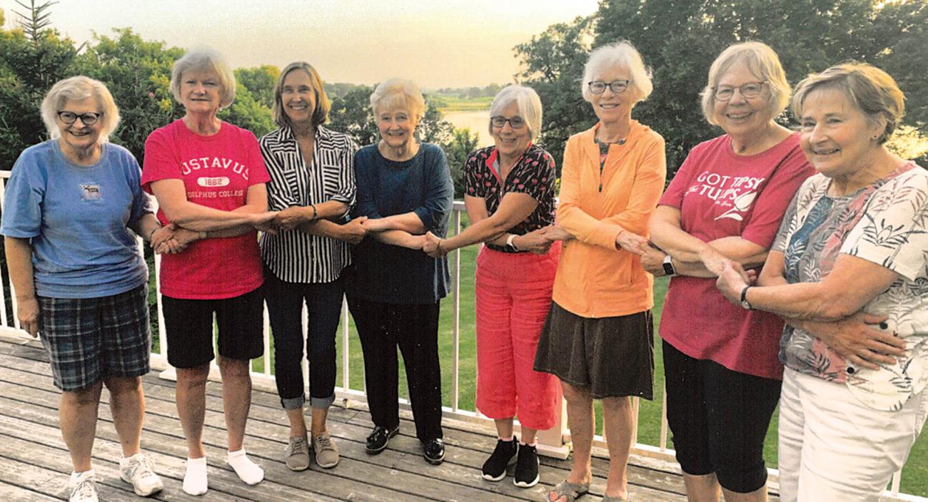 Eight mature women hold hands near a lake. 