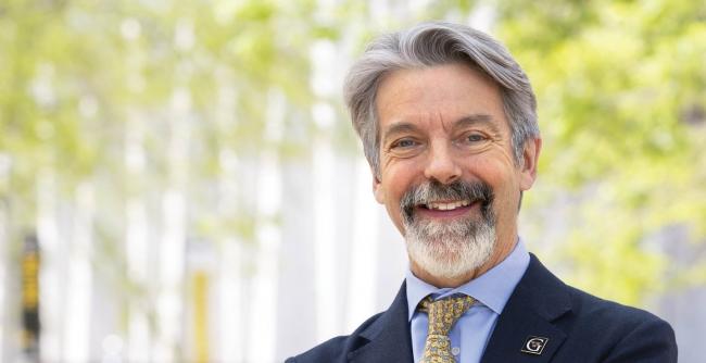 A distinguished gentleman stands on a college campus, smiling, wearing a pin with the "G" logo of Gustavus Adolphus College. 