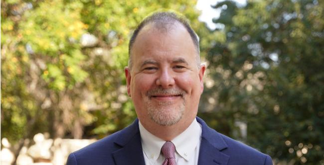 A distinguished gentleman stands on a college campus, smiling, wearing a pin with the "G" logo of Gustavus Adolphus College. 