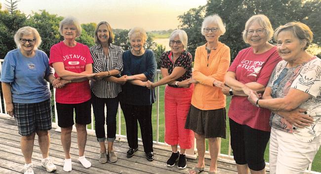 Eight mature women hold hands near a lake. 