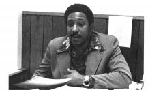 A 1970s era black man with a mustache and cool pointy shirt collar gestures while speaking. 