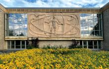 The bas relief sculpture reminiscent of DaVinci's Vetruvian Man on the side of the Nobel Hall of Science, with a bed of prairie flowers and blue sky and clouds reflected in the background. 