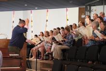 A man in a samurai hairstyle conducts a choir in a church balcony.