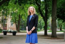 A woman professor stands on a college campus, smiling at the camera. 