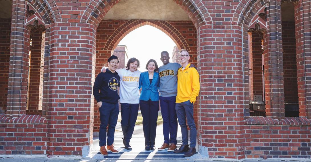 A woman stands surrounded by young adults in Gustavus Adolphus College gear. 