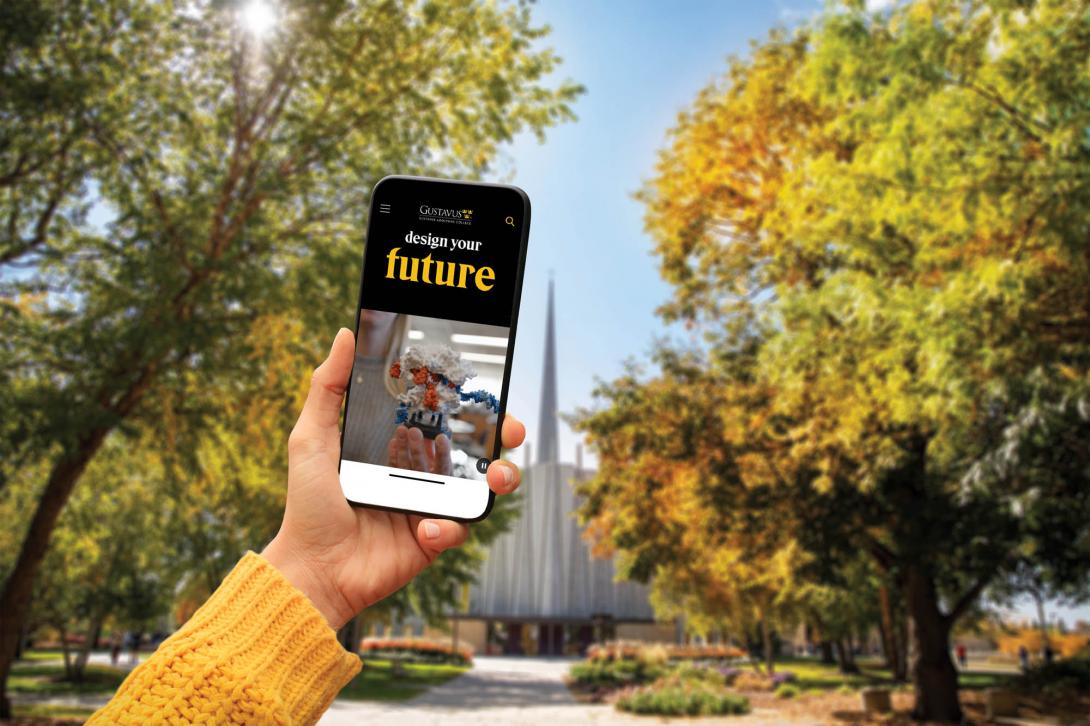A hand holds up a phone with the Gustavus website on it, on a college campus in the fall. 