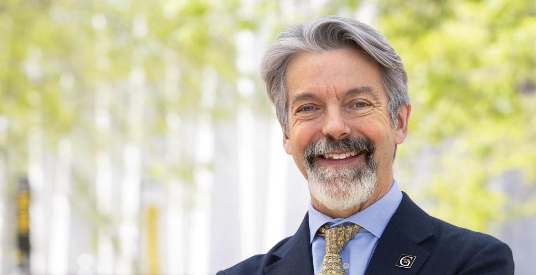 A distinguished gentleman stands on a college campus, smiling, wearing a pin with the "G" logo of Gustavus Adolphus College. 