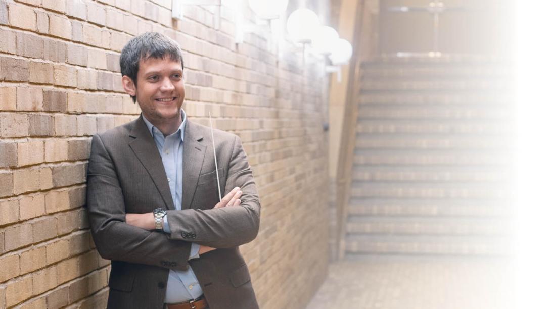 A man with a conductor's baton leans against a brick wall in a hallway. 