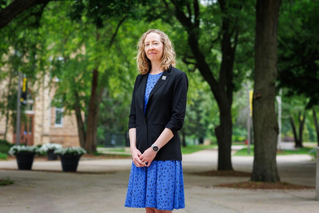 A woman professor stands on a college campus, smiling at the camera. 
